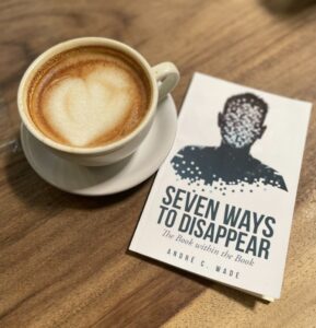 Seven Ways to Disappear, a book by Andre C. Wade. Pictured next to a cup of coffee on a wooden table, the book shows a white cover with a pixelated graphic of a person, suggesting their disappearing act.
