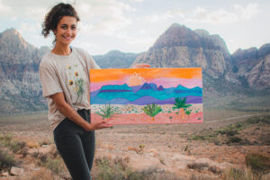 Michelle LaFiura, The Lost Journalist, with her artwork, a colorful painting of the mountains, in front of her inspiration at Red Rock Canyon.