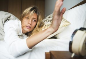 Woman laying in bed reaching over to turn off or snooze her clock, representing Be Late For Something Day.