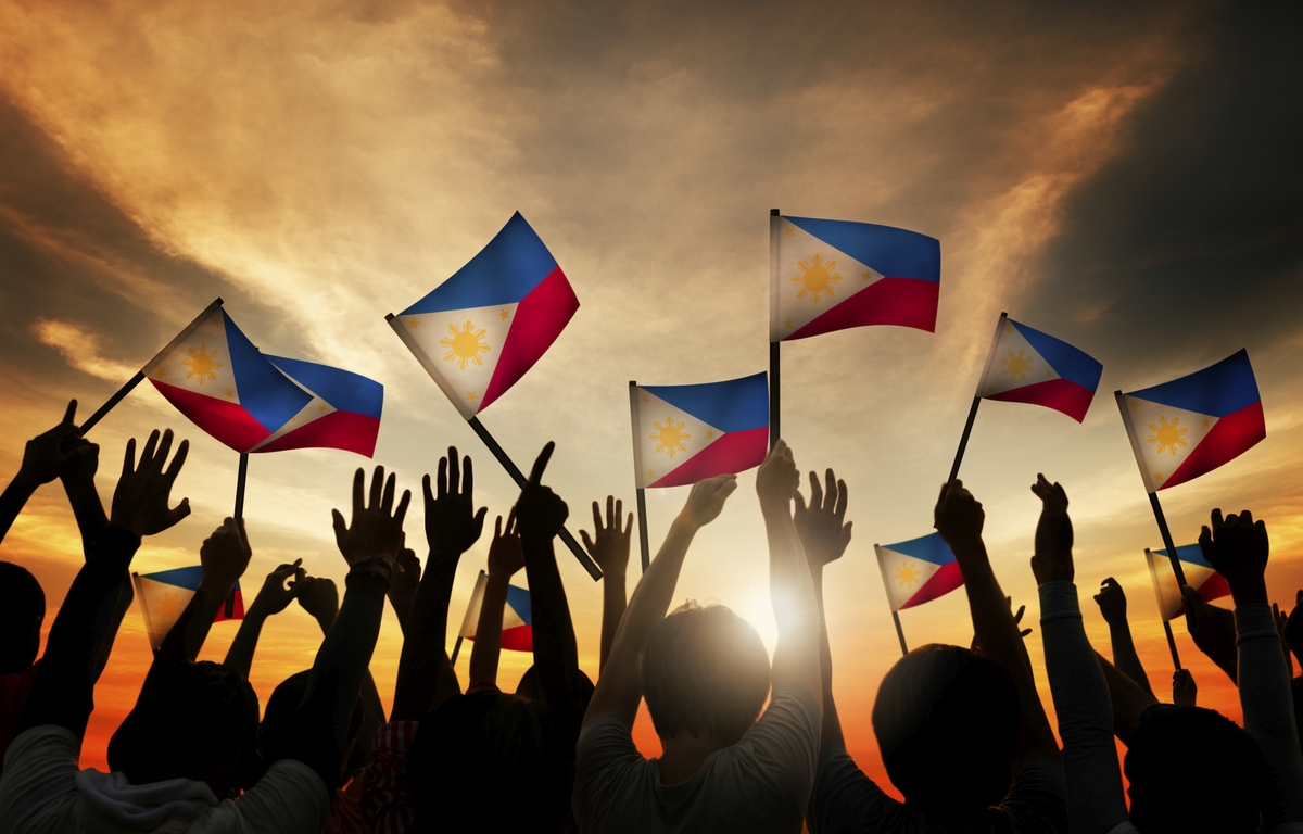 Group of people waving Filipino flags over a backlit sunset sky