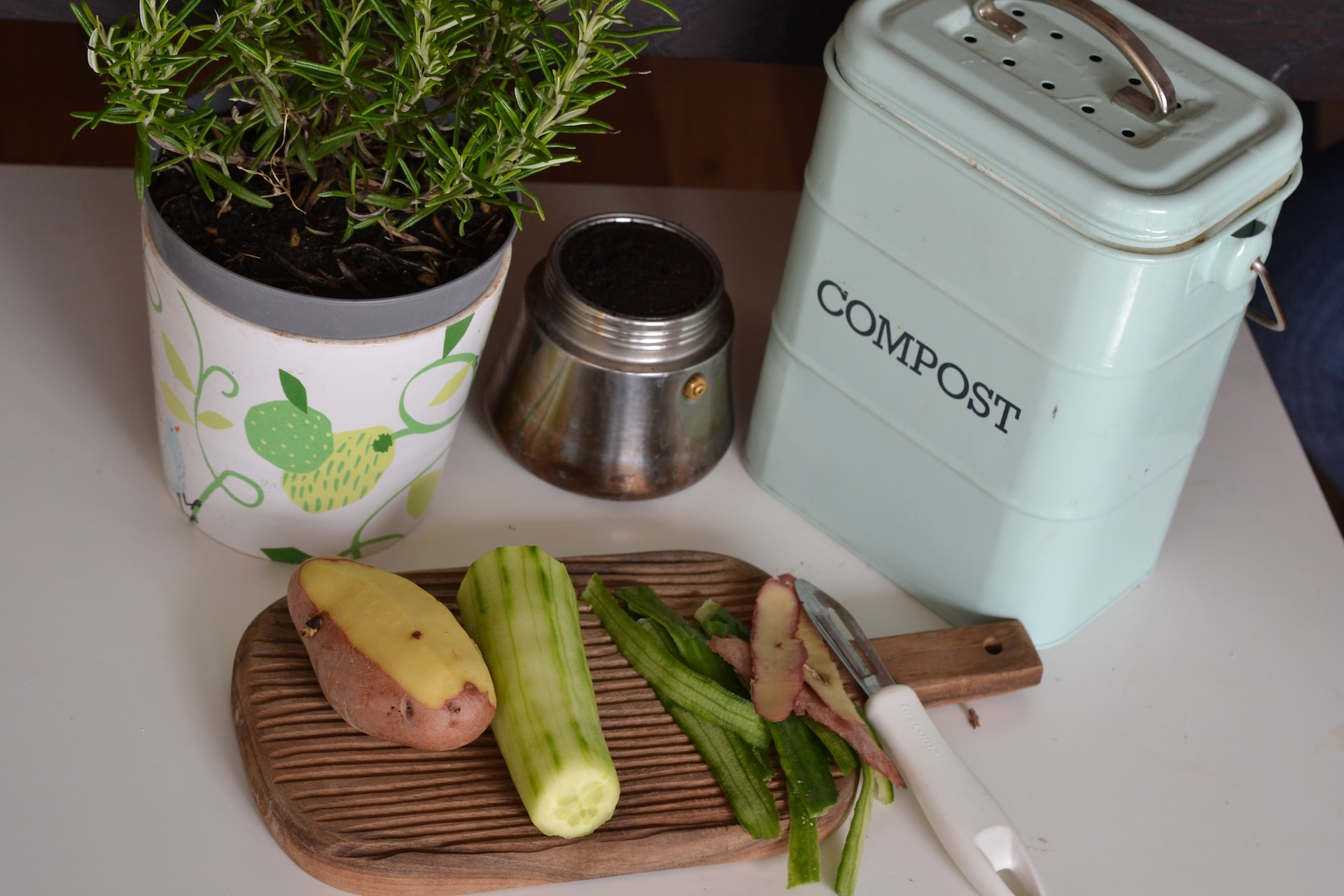 Picture of a small cutting board with a potato and cucumber being peeled. A small can that says 'Compost' is behind that to the right, and a small pot of rosemary is behind to the left.